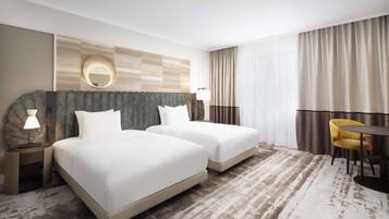 Ein Hotelzimmer mit zwei Betten, einem runden Spiegel, einem Schreibtisch mit Lampe, einem Stuhl und einem großen Fenster mit Vorhängen.