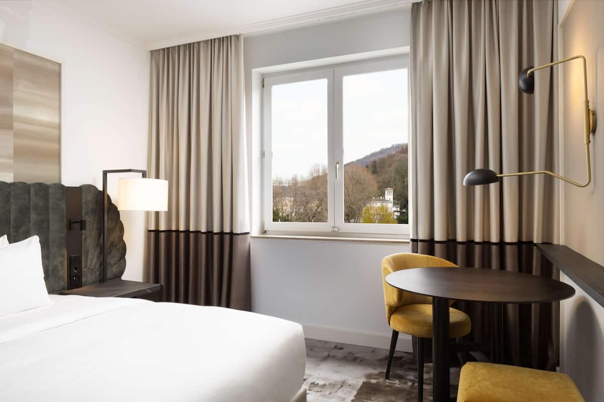 A hotel room with a bed, a desk with a lamp, a chair, a small table, and a view of the outdoors through a window.