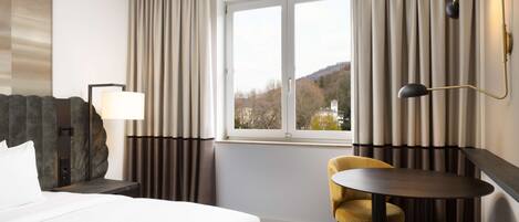 A hotel room with a bed, a desk with a lamp, a chair, a small table, and a view of the outdoors through a window.