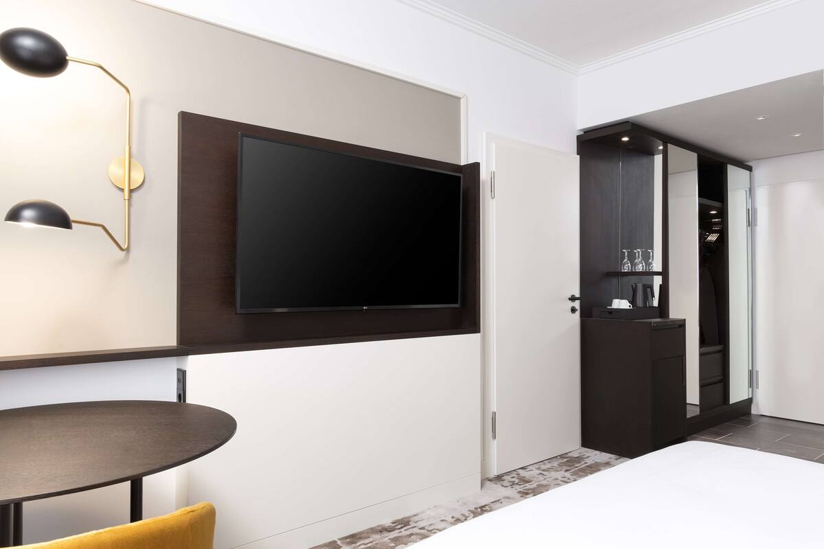 A modern hotel room with a large flat-screen TV mounted on the wall, a round dining table with a yellow chair, and a dark wooden cabinet with a mirror.