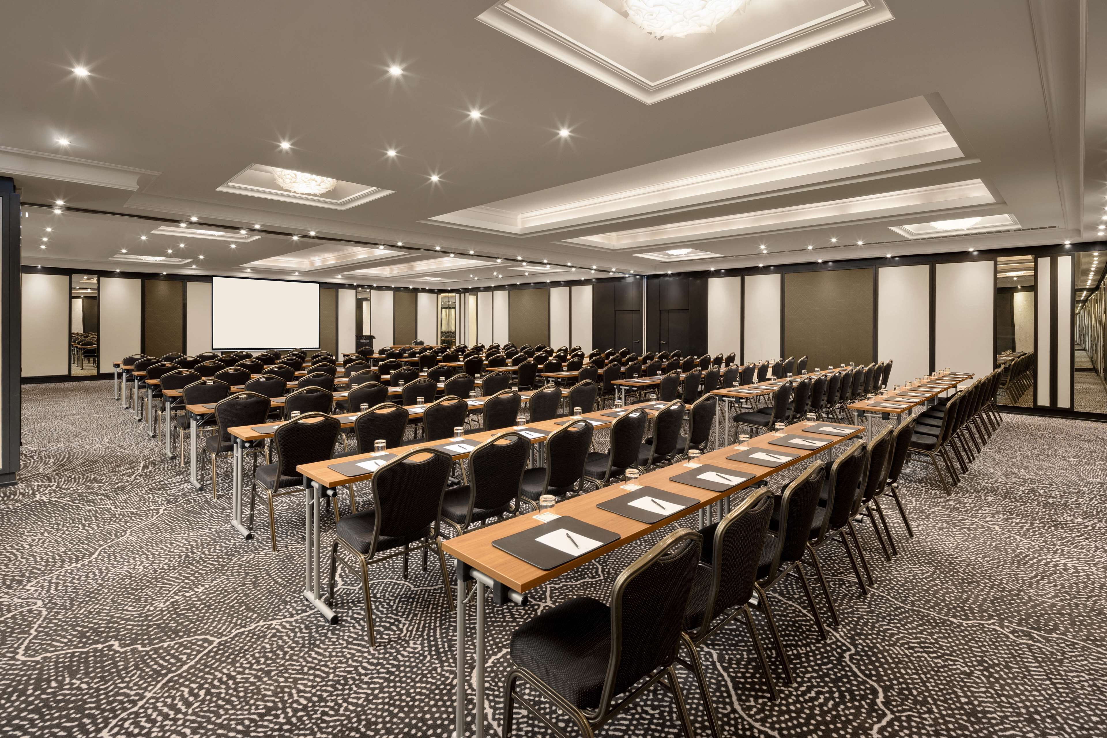 A conference room with rows of tables and chairs, a projector screen, and a carpeted floor.