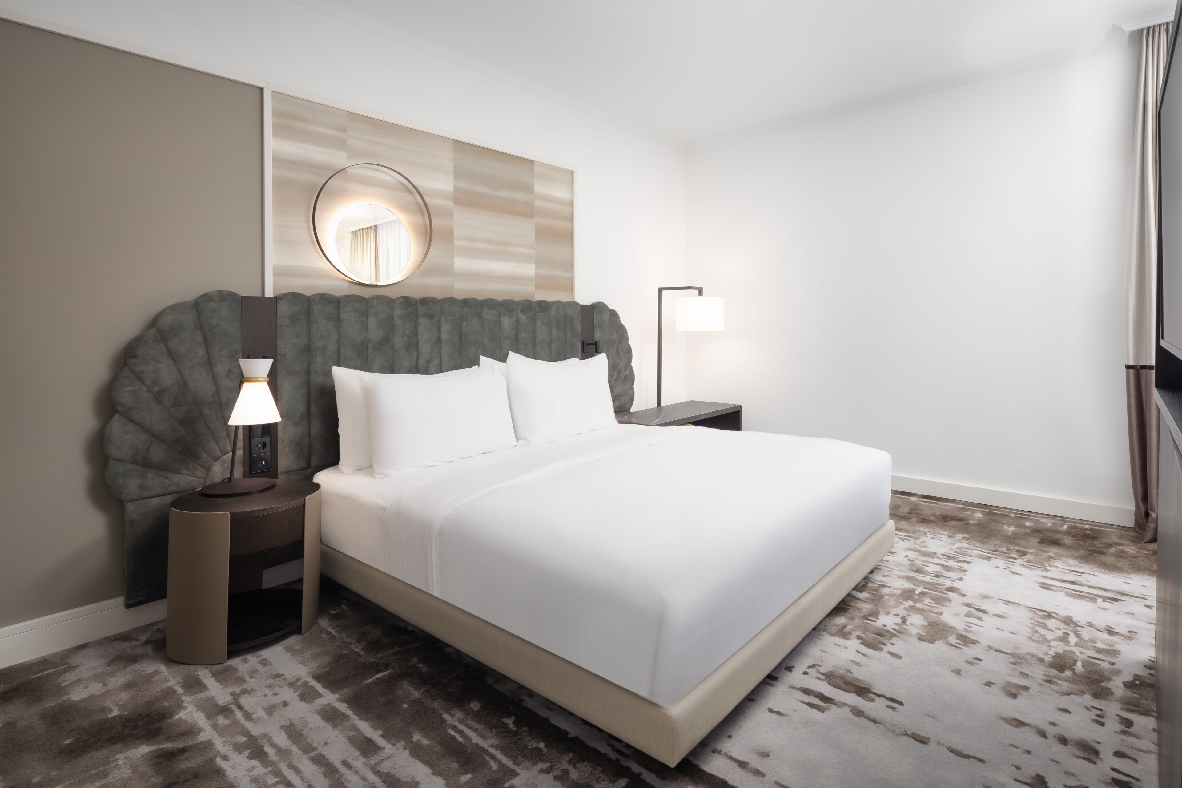 A neatly made bed with white linens, a round mirror above the headboard, and a nightstand with a lamp.