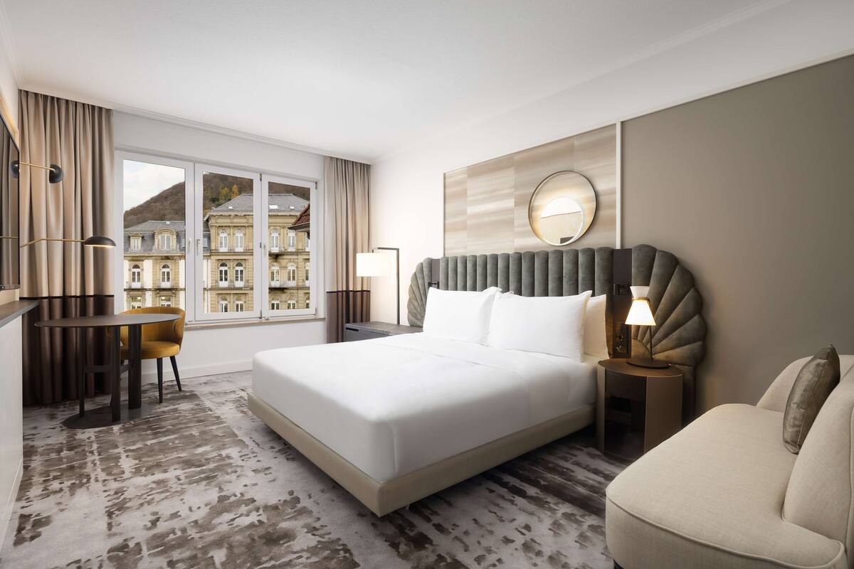 A hotel room with a large bed, a desk, a chair, a round mirror, and a view of buildings through the window.