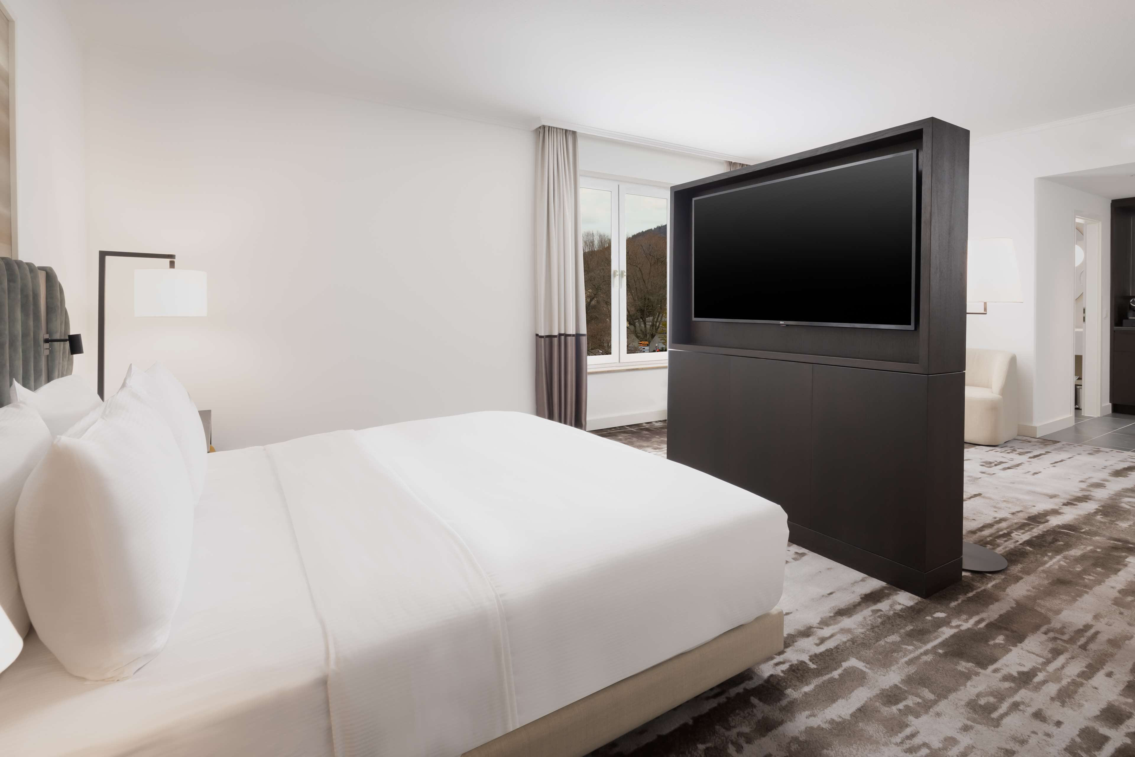 Ein Hotelzimmer mit einem großen Bett, einem Flachbildfernseher, einem Schreibtisch und Blick ins Freie.