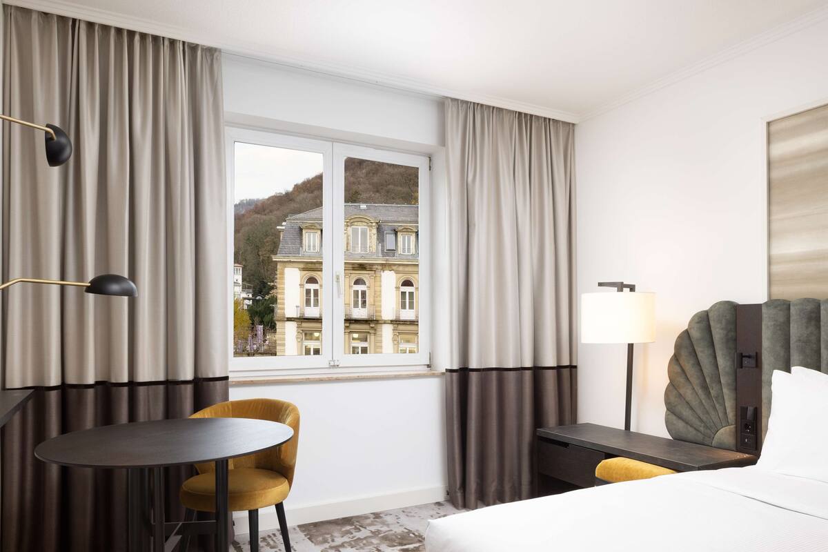 A hotel room with a bed, a desk, a chair, a lamp, and a view of a building through a window.