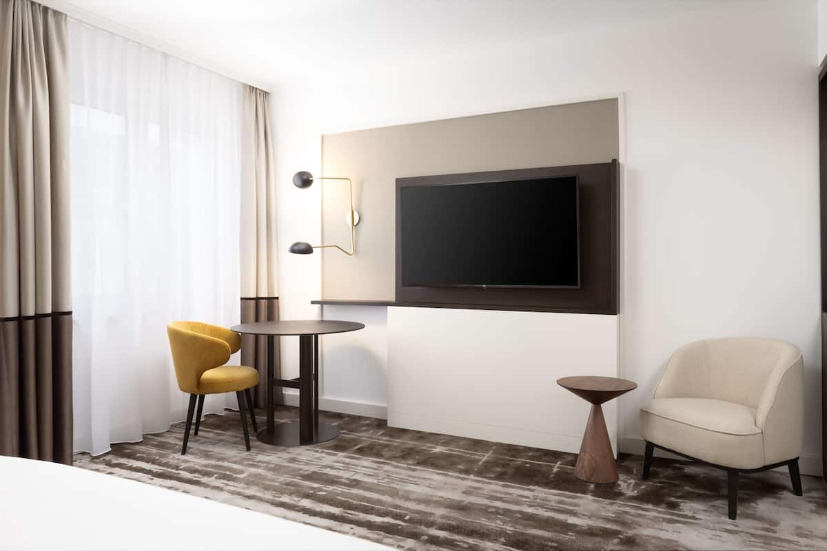 A hotel room with a large flat-screen TV mounted on the wall, a round table with two chairs, a small wooden side table, and a cream-colored armchair.