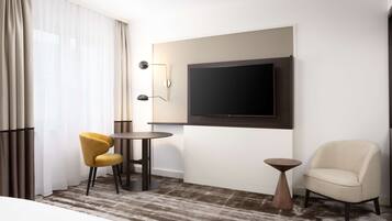 Ein Hotelzimmer mit einem großen Flachbildfernseher an der Wand, einem runden Tisch mit zwei Stühlen, einem kleinen Nachttisch und einem cremefarbenen Sessel.
