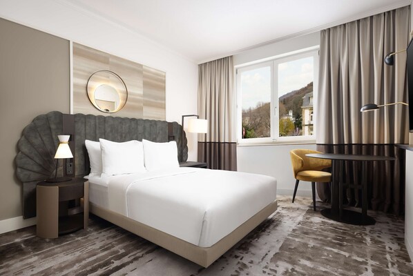 Habitación de hotel con una cama grande, lámparas de noche, un espejo redondo, un escritorio con silla y vistas al exterior.