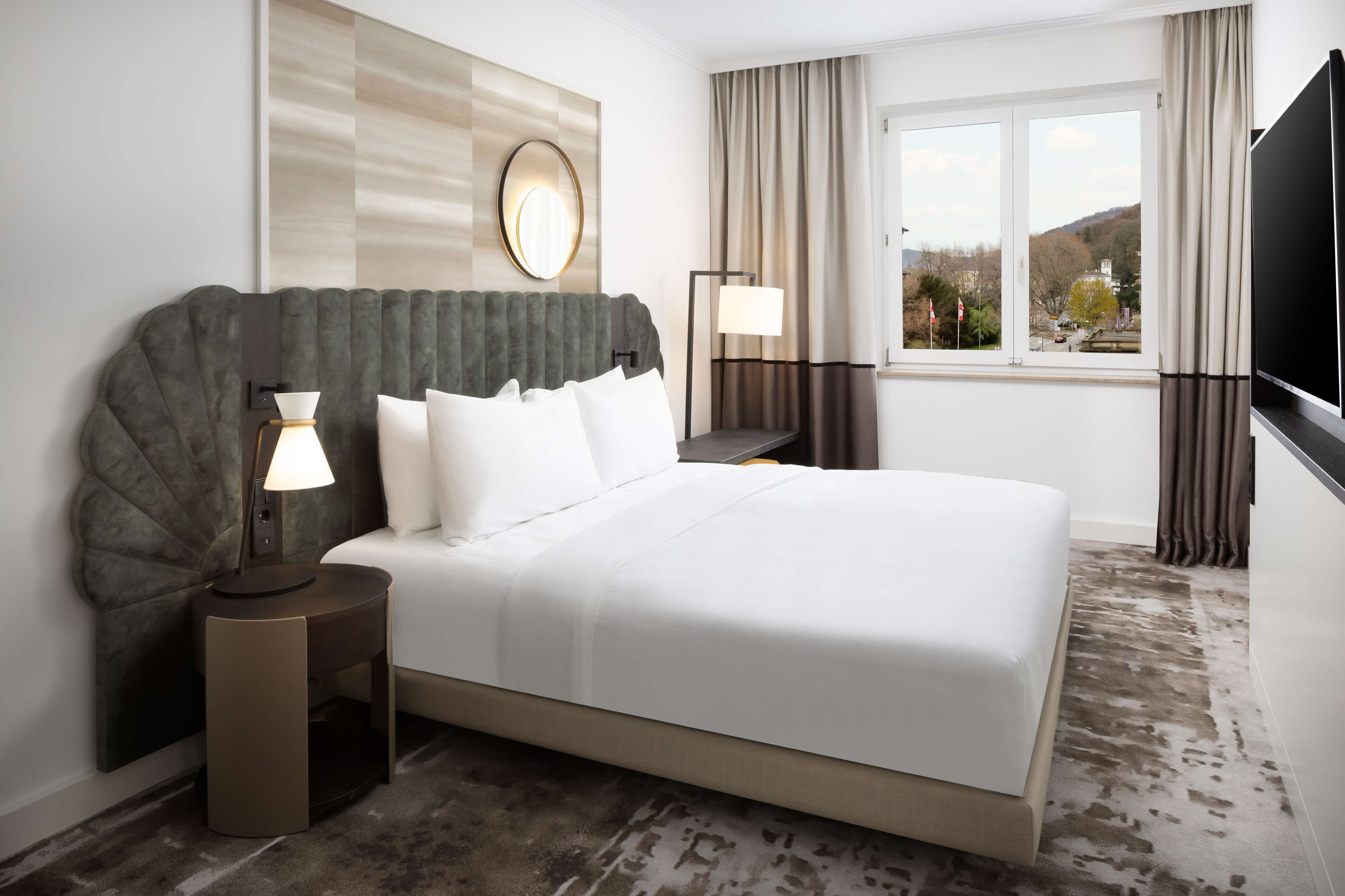 Una habitación de hotel moderna con una cama grande, una mesita de noche con lámpara, un espejo en la pared y una ventana con cortinas.