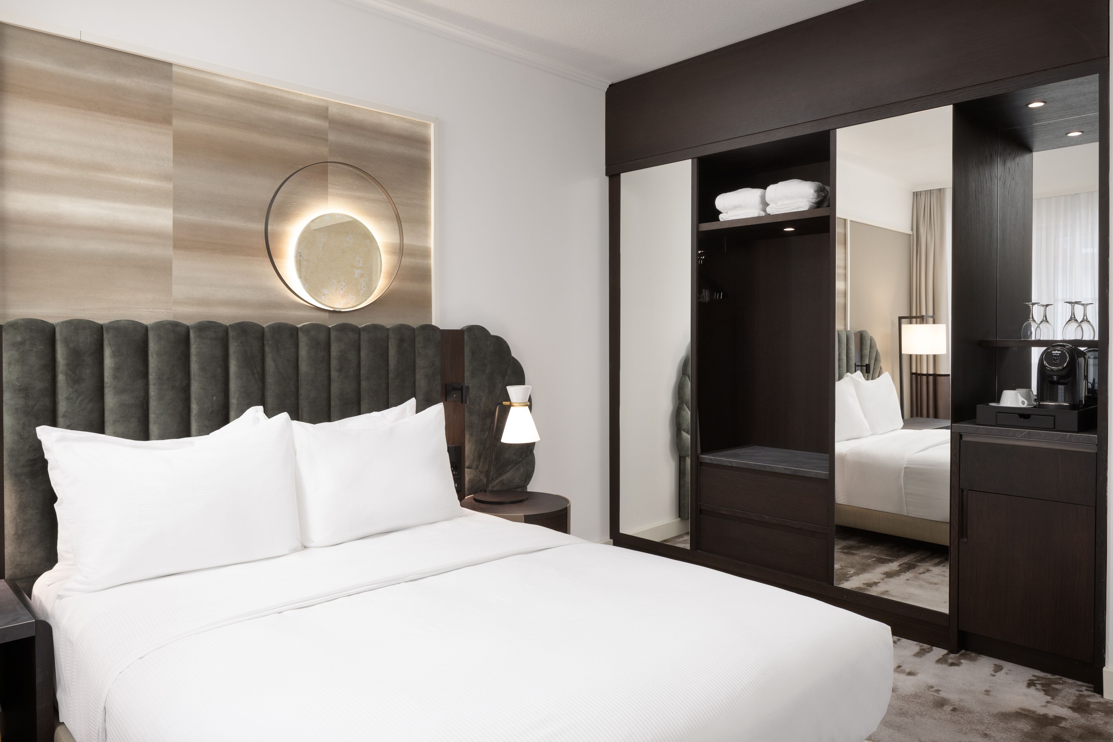 A hotel room with a large bed, a round mirror, a wardrobe, and a nightstand with a lamp.
