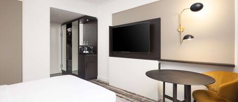 A hotel room with a bed, a small round table, a yellow chair, a TV mounted on the wall, and a kitchenette in the background.