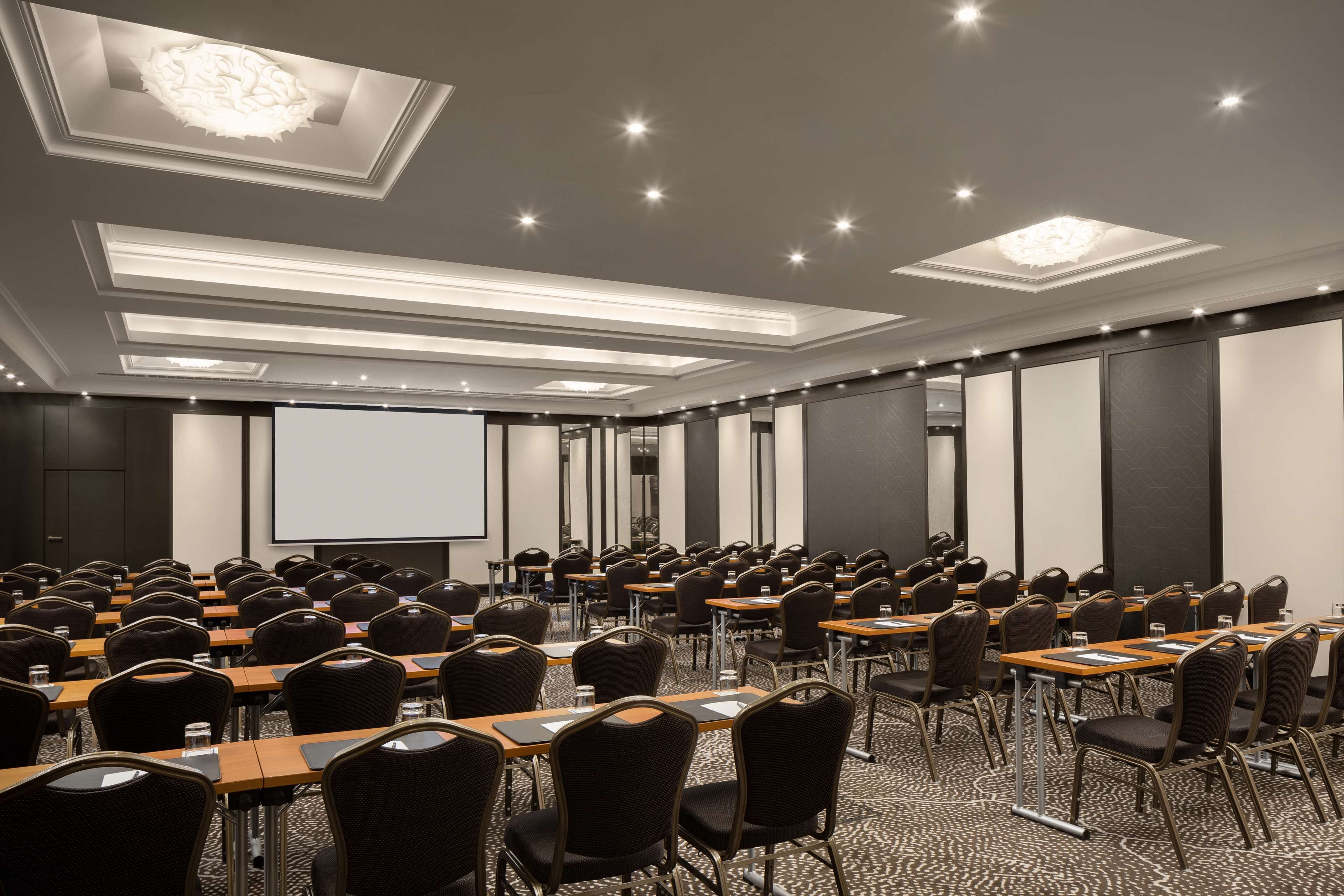 Un salón de conferencias con filas de mesas y sillas, una pantalla de proyector e iluminación empotrada en el techo.