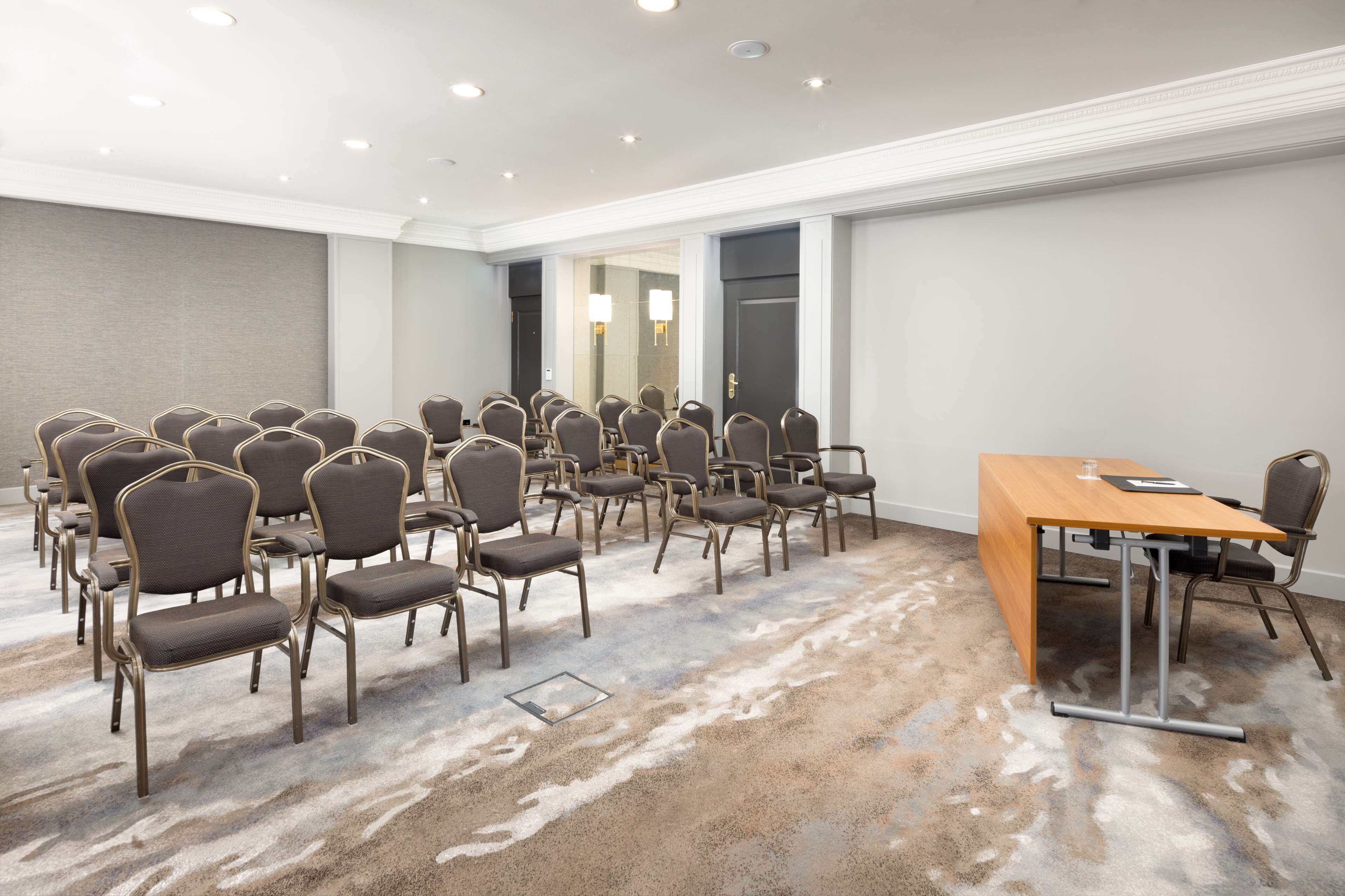 Sala de conferências com fileiras de cadeiras e uma mesa, piso coberto de carpete e luminárias embutidas no teto.