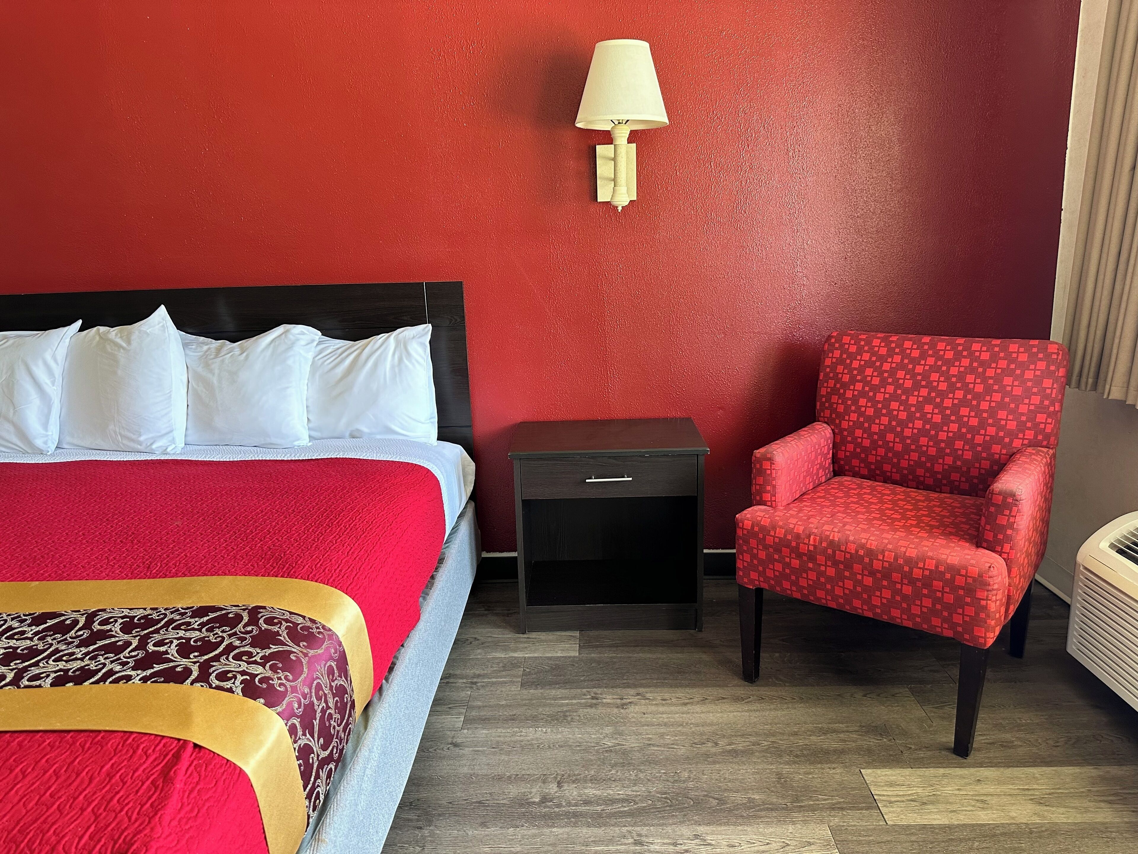 A hotel room with a bed, a red armchair, a nightstand, and a wall-mounted lamp.