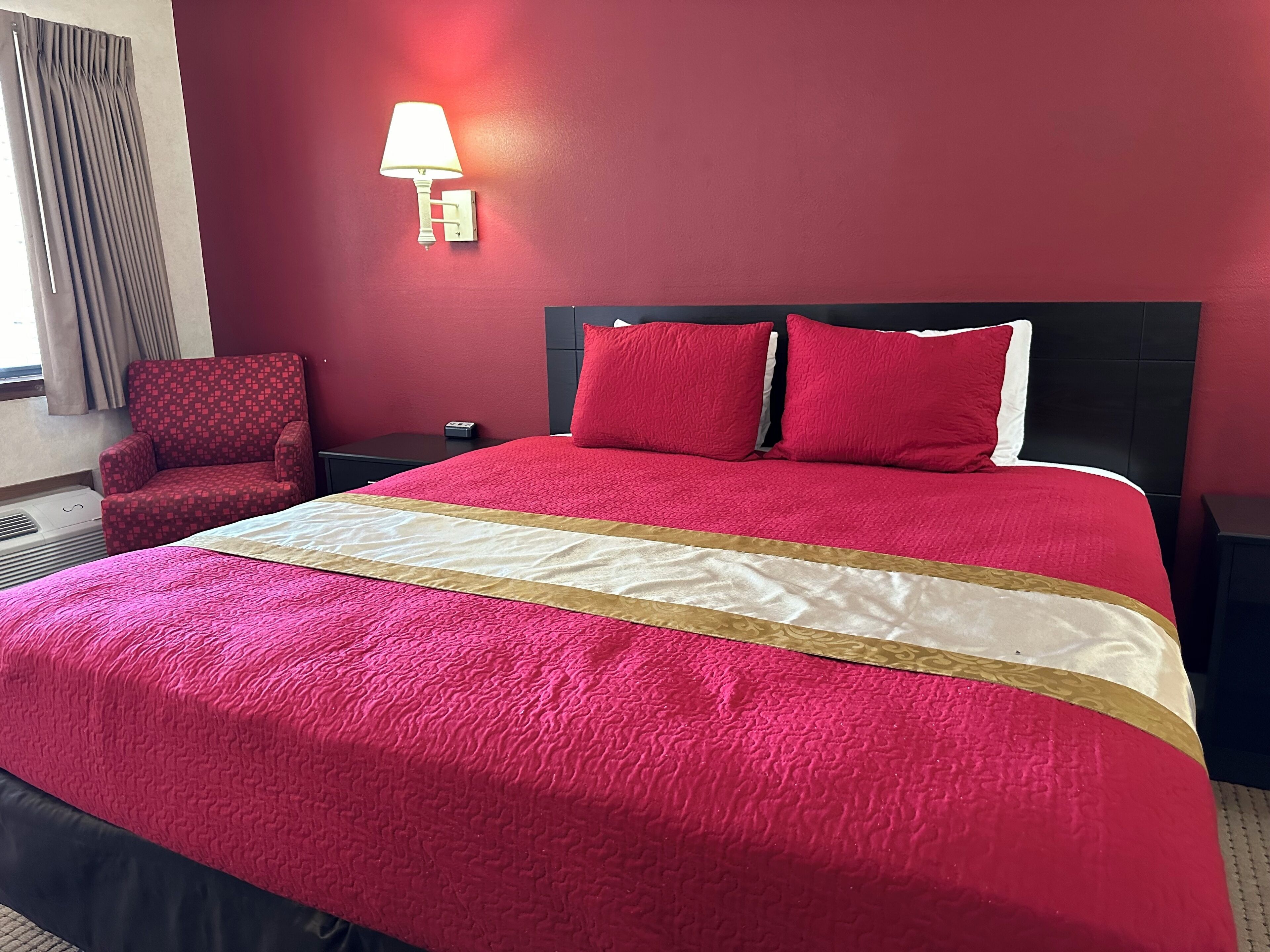 A hotel room with a large bed, red walls, a red armchair, a nightstand, and a lamp.