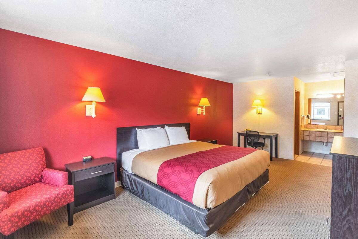 A hotel room with a red wall, a bed with a beige and red bedspread, a red armchair, a small desk with a lamp, and a bathroom visible through an open door.