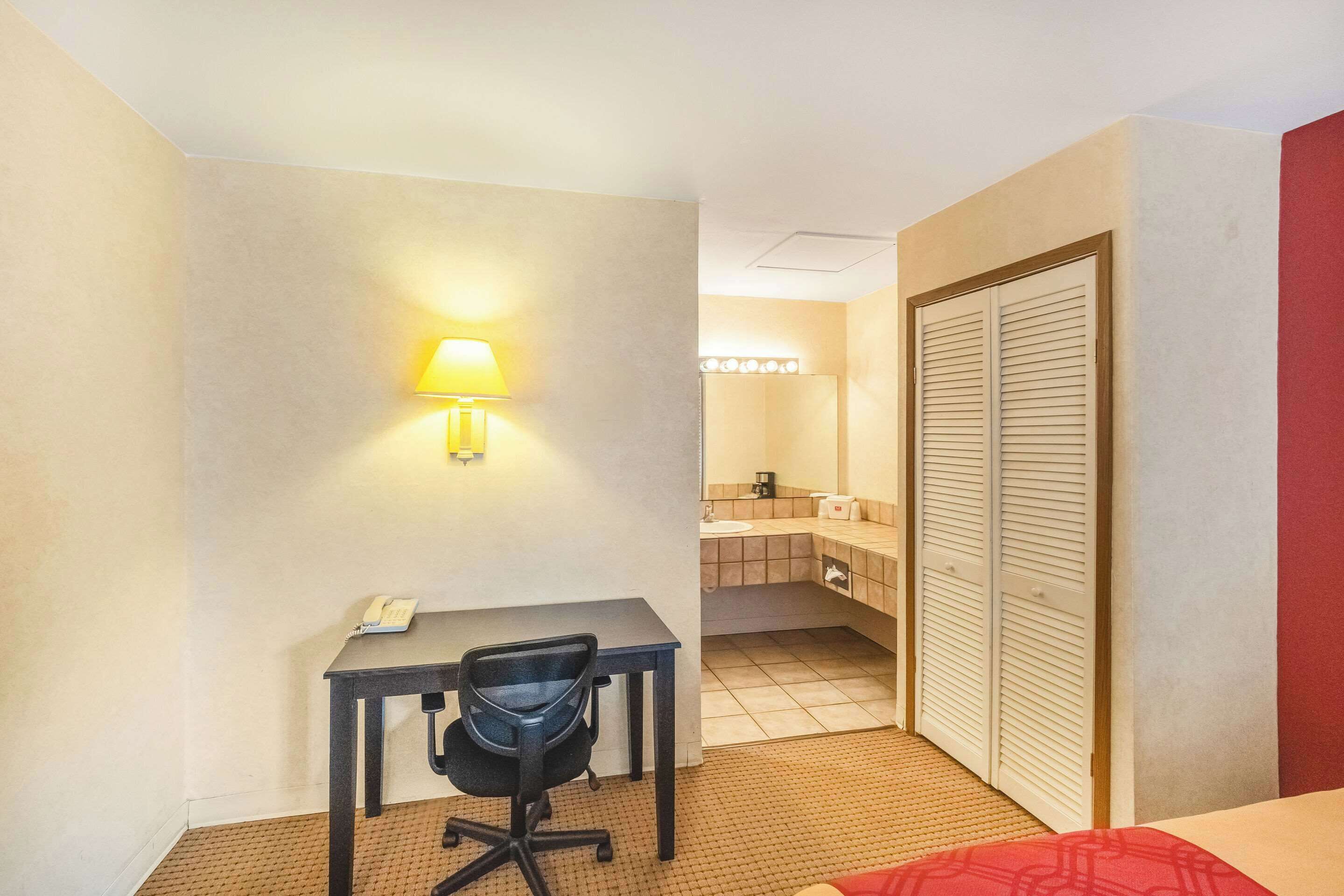 A hotel room with a desk, chair, bed, and a bathroom with a mirror and sink.