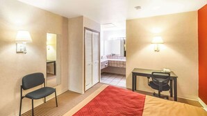 Une chambre d’hôtel avec un lit, un bureau, une chaise et une salle de bain visible à travers une porte ouverte.