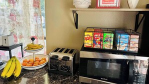 Un comptoir de cuisine avec un grille-pain, un four à micro-ondes, un bol d’oranges, une assiette de bananes et une étagère remplie de boîtes de céréales.