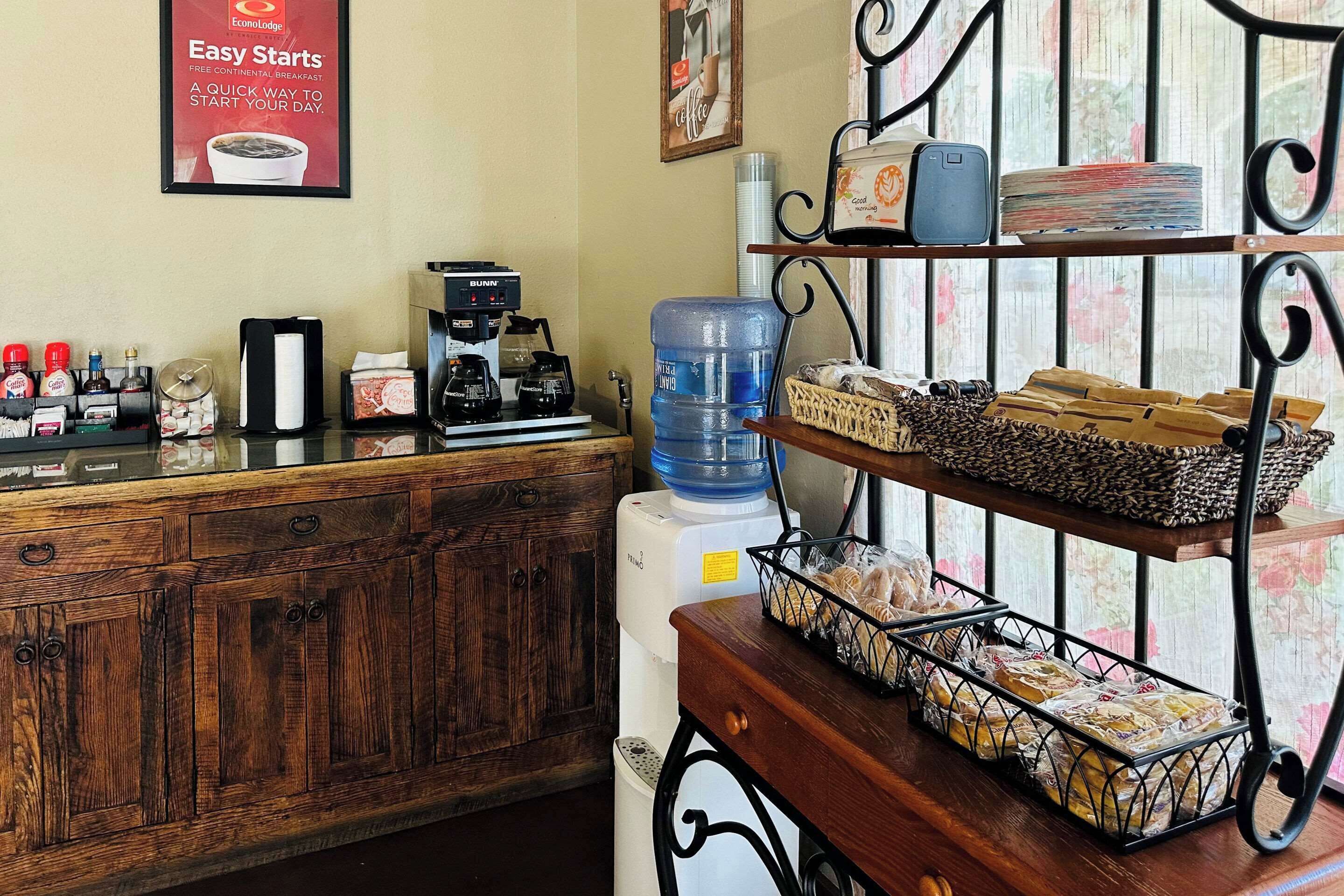 Zona de recepción con mostrador de madera, máquinas de café, dispensador de agua y cestas de pan en estantes.