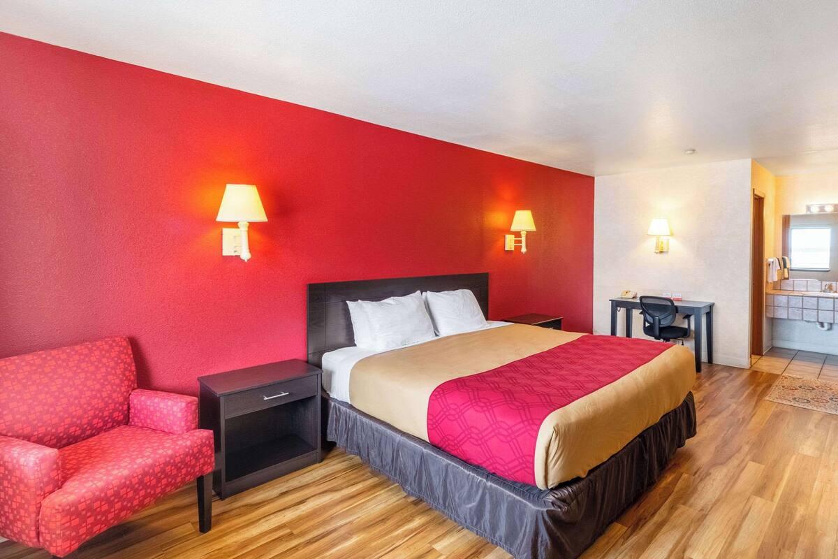 A hotel room with a red wall, a bed with a beige and red bedspread, a red armchair, a nightstand, a desk with a chair, and a small window.