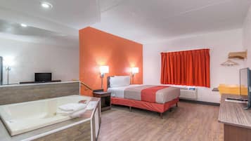 A hotel room with a large bathtub, a bed with red bedding, a bedside table with a lamp, and a window with red curtains.