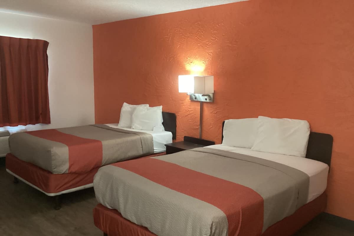 A hotel room with two beds, each with a red and beige bedspread, white pillows, and a nightstand with a lamp.