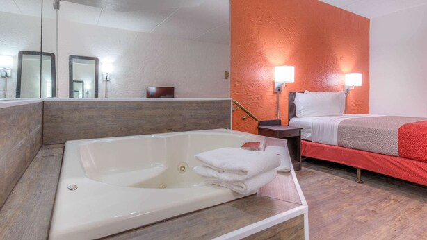 A hotel room with a large bathtub, a bed with red bedding, and a bright orange accent wall. - Motel 6 Bridgeview, IL (Bridgeview)