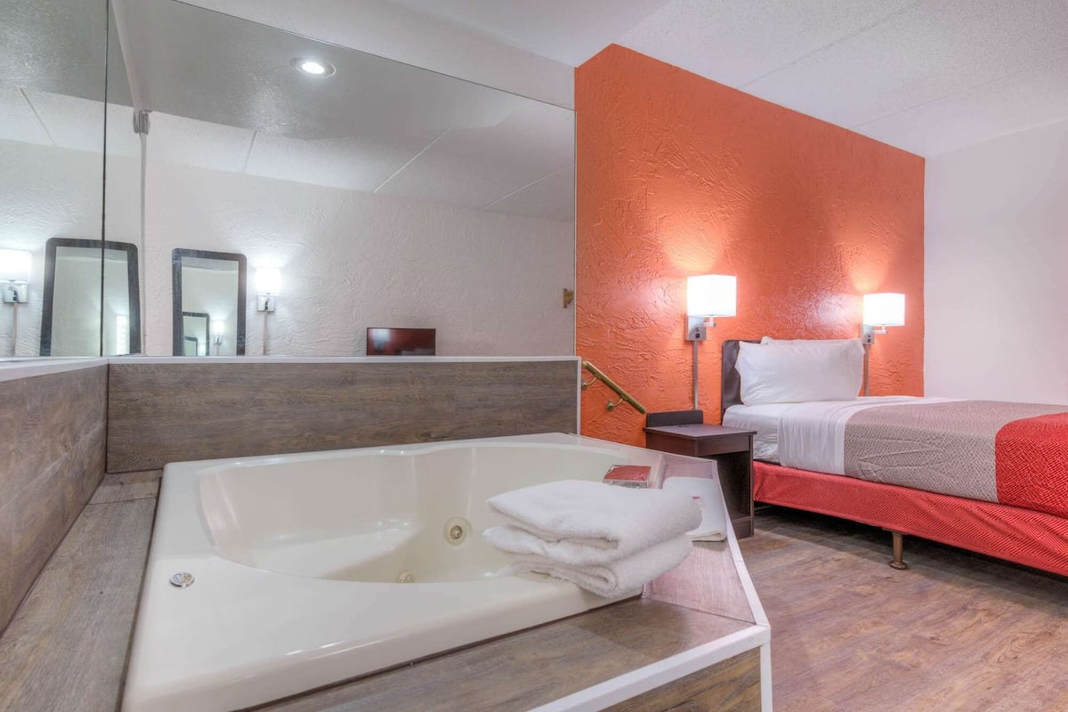 A hotel room with a large bathtub, a bed with red bedding, and a bright orange accent wall.