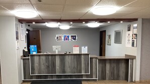 A reception desk with a wooden finish, a black mat in front, and a tiled floor. There are three ceiling lights, a door to the left, and a wall with framed pictures and signs. - Motel 6 Bridgeview, IL (Bridgeview)