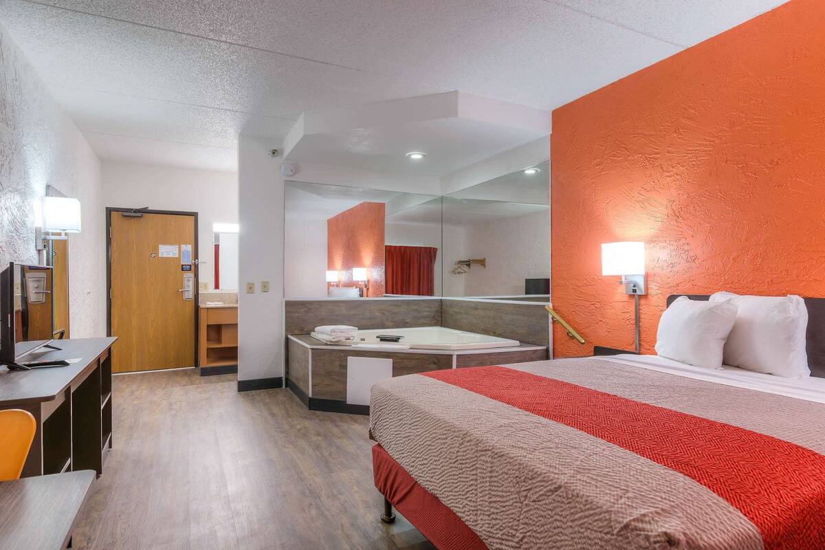 A hotel room with a bed, desk, chair, and a bathroom visible through a glass partition.