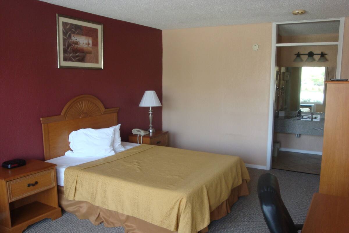 A hotel room with a bed, a nightstand, a lamp, a chair, and a bathroom visible through an open door.