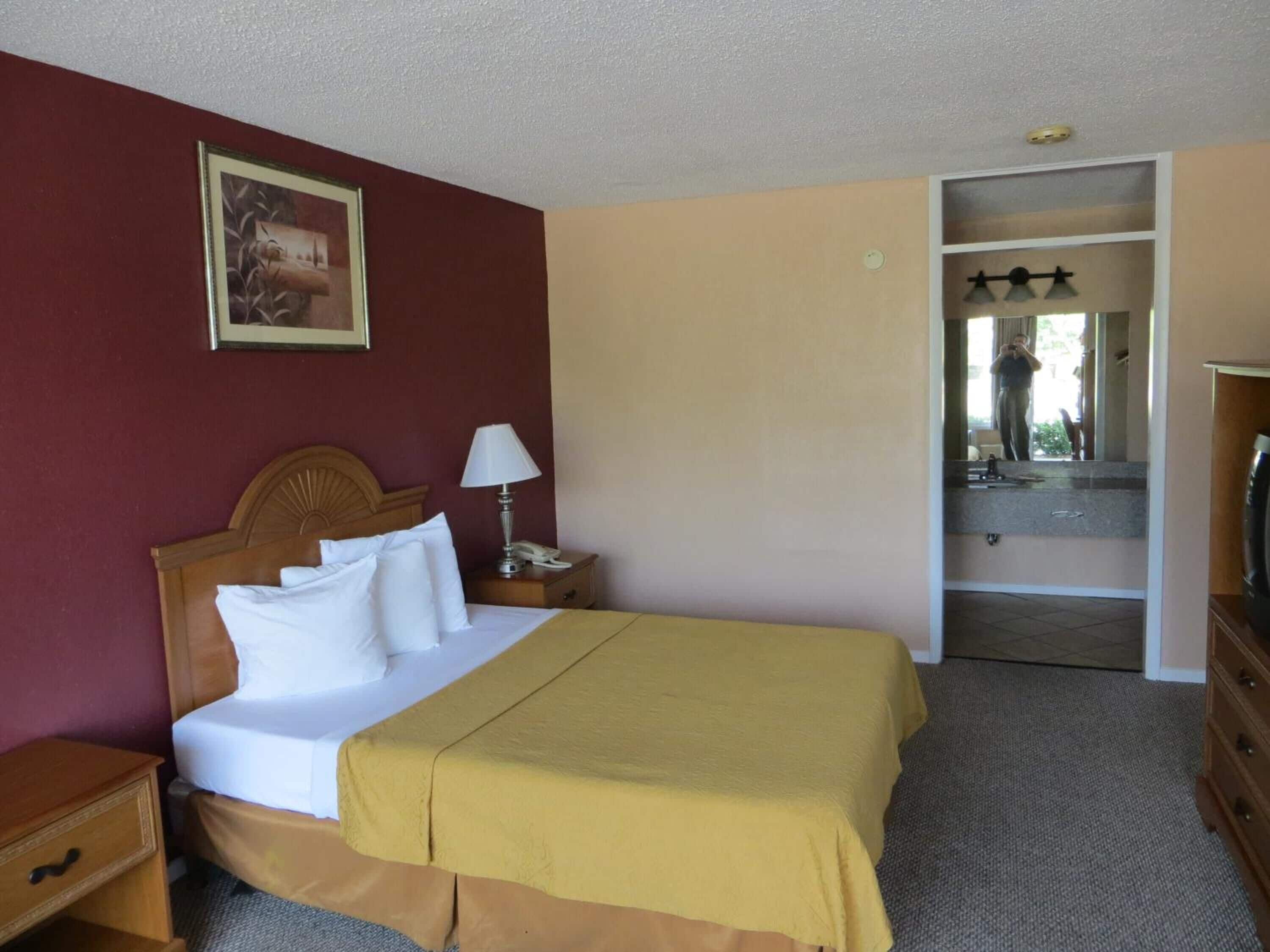 A hotel room with a bed, a nightstand, a lamp, a painting on the wall, and a mirror reflecting a person taking a photo.