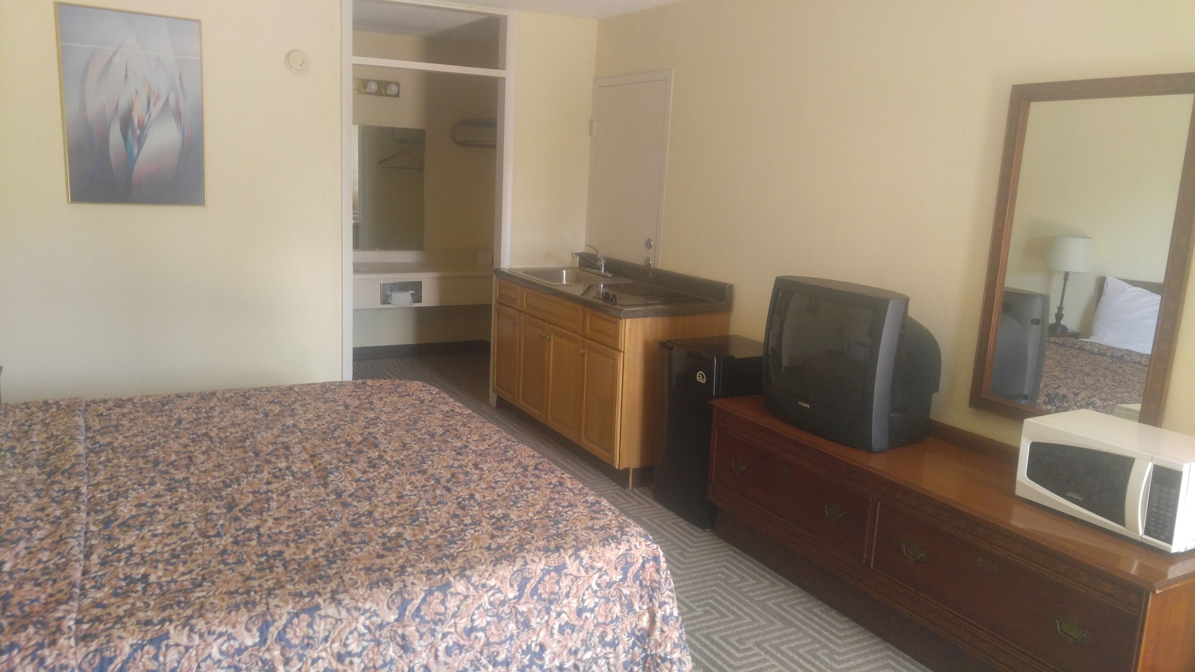 A hotel room with a bed, a wooden dresser, a television, a microwave, and a bathroom visible through an open door.