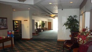 A hotel lobby with a patterned carpet, a reception desk, a refrigerator, and a seating area with a red chair and a potted plant.