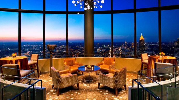 A high-rise hotel room with a panoramic city view, a round table with chairs, and a modern chandelier.