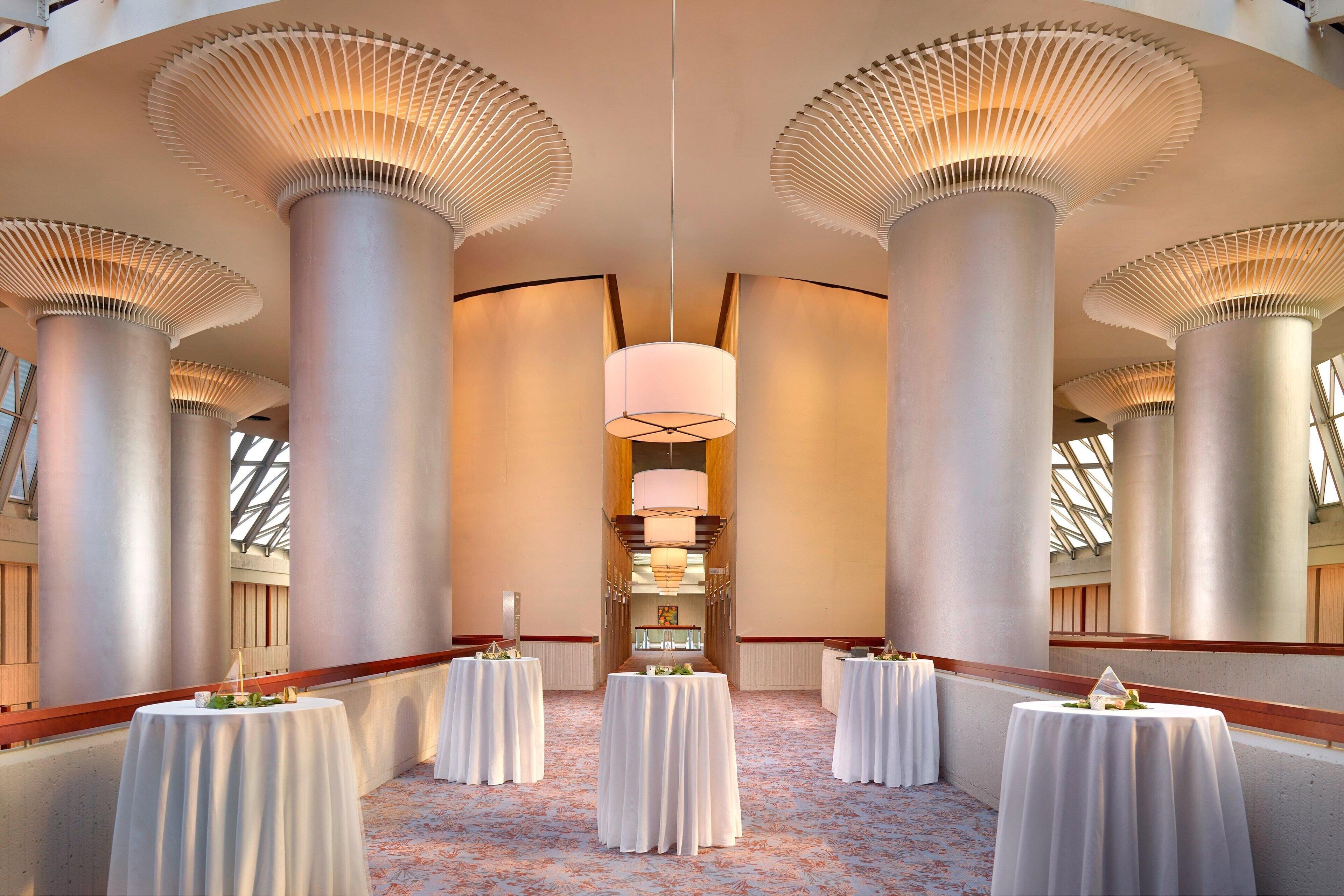 A hotel corridor with high ceilings, large columns, and round tables with white tablecloths.