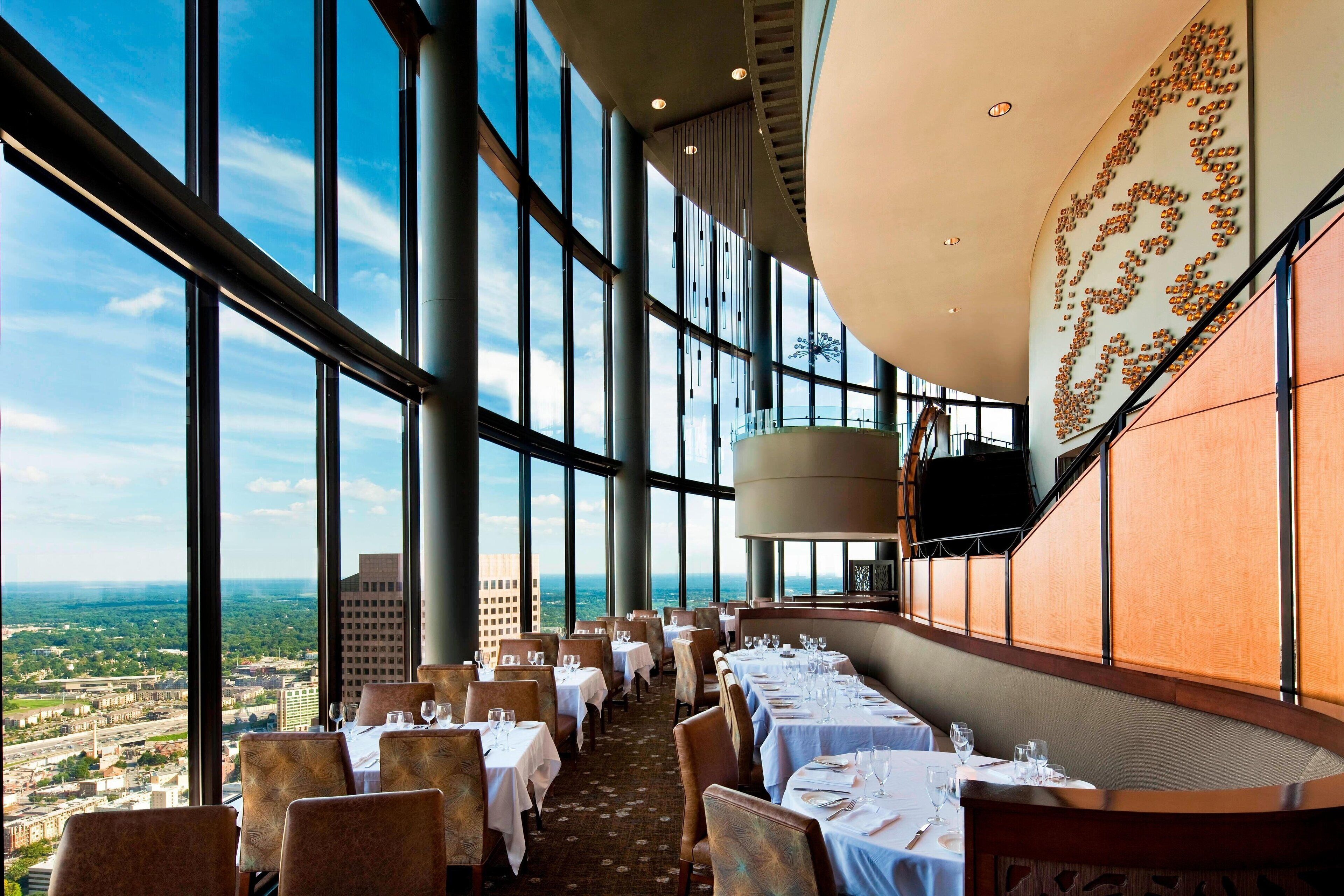 A high-ceiling restaurant with large windows offering a city view. Tables are set with white tablecloths and chairs.