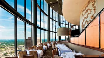 Un restaurant avec de hauts plafonds et de grandes fenêtres offrant une vue sur la ville. Les tables sont dressées avec des nappes blanches et des chaises.