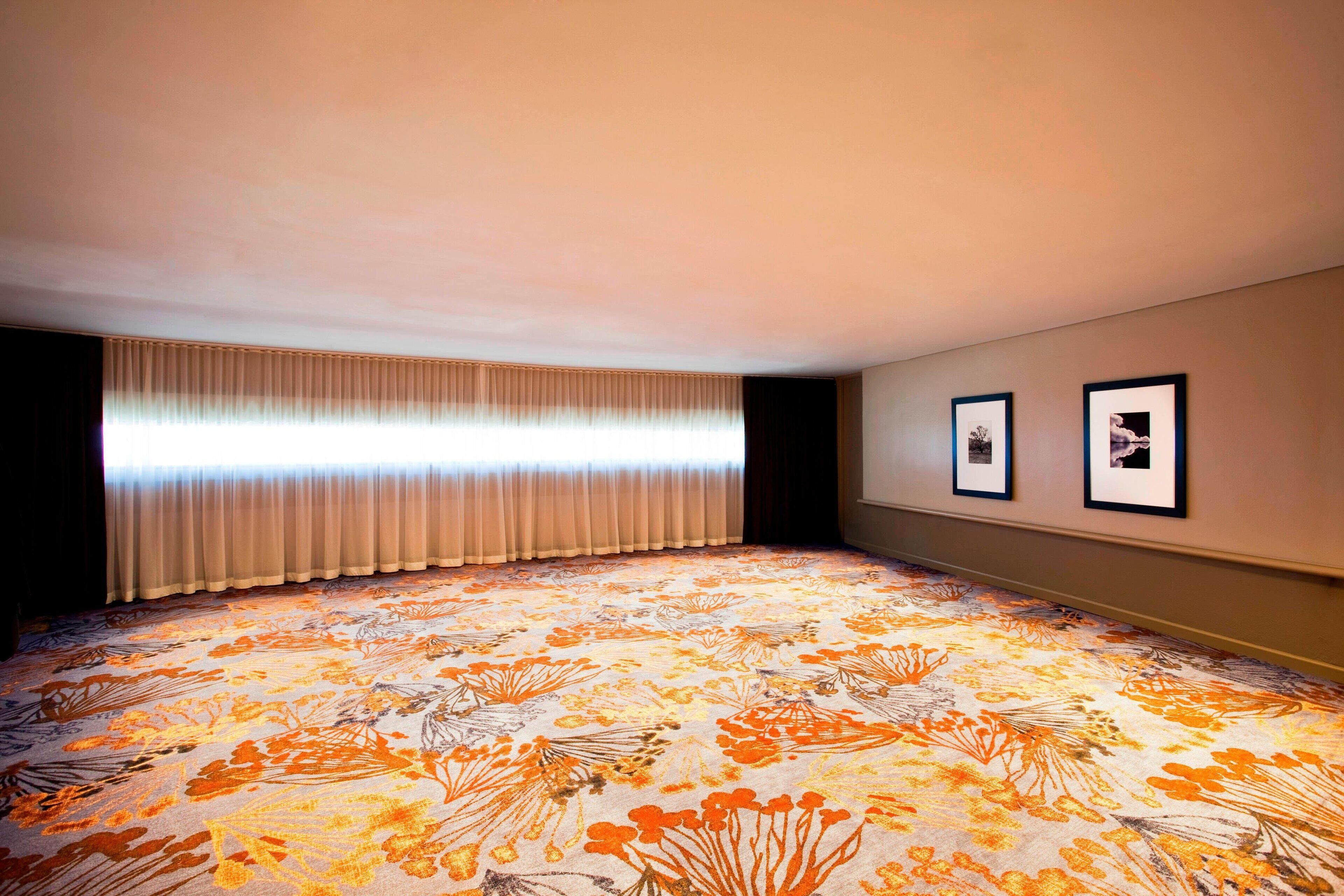 A room with a patterned carpet, a large window with curtains, and two framed pictures on the wall.