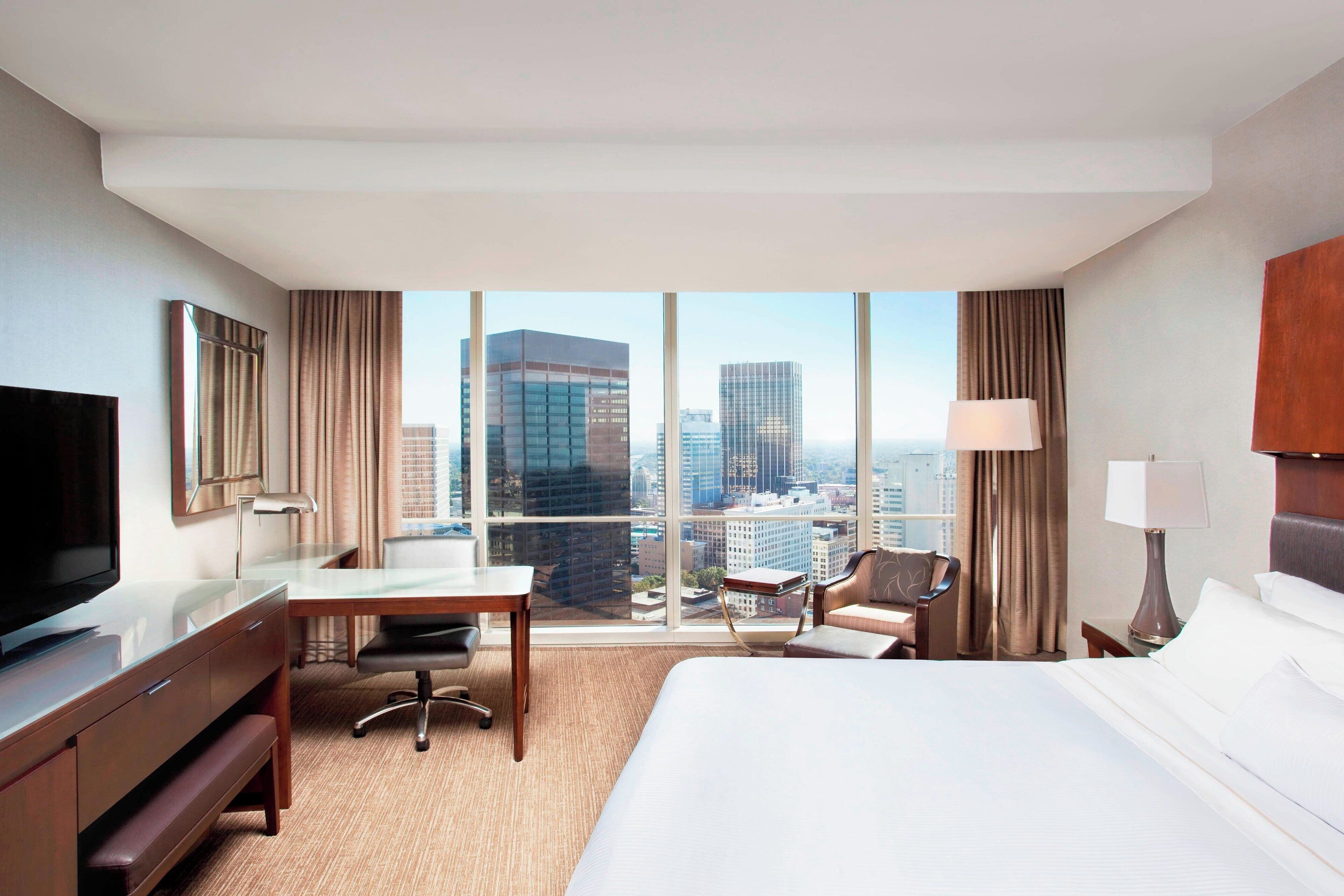 Quarto de hotel com uma cama grande, uma mesa, uma cadeira, uma TV e vista para a paisagem urbana através de grandes janelas.