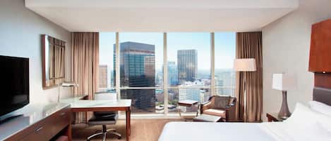 Quarto de hotel com uma cama grande, uma mesa, uma cadeira, uma TV e vista para a paisagem urbana através de grandes janelas.