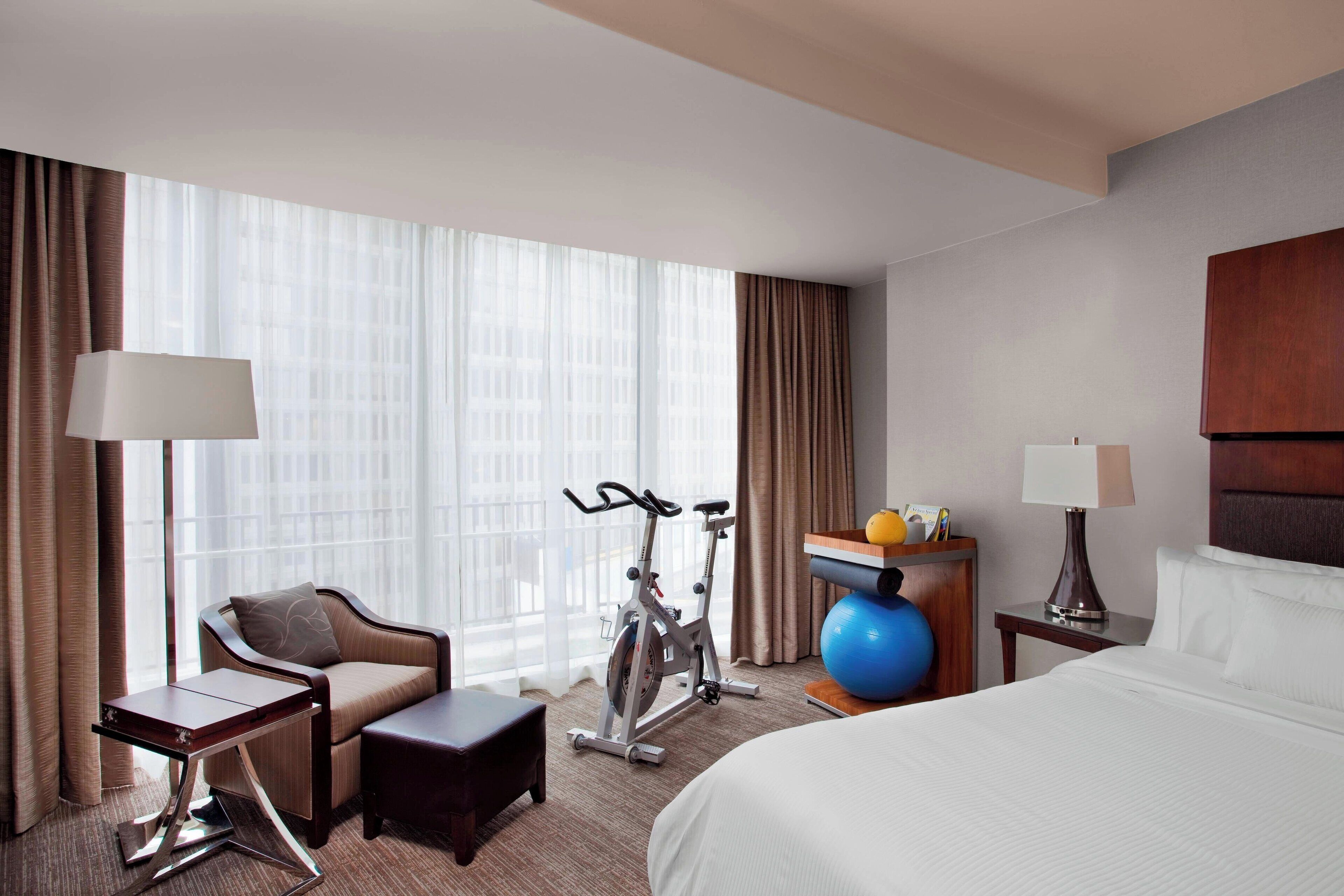 A hotel room with a bed, a desk, a chair, an exercise bike, a small table, and a window with curtains.