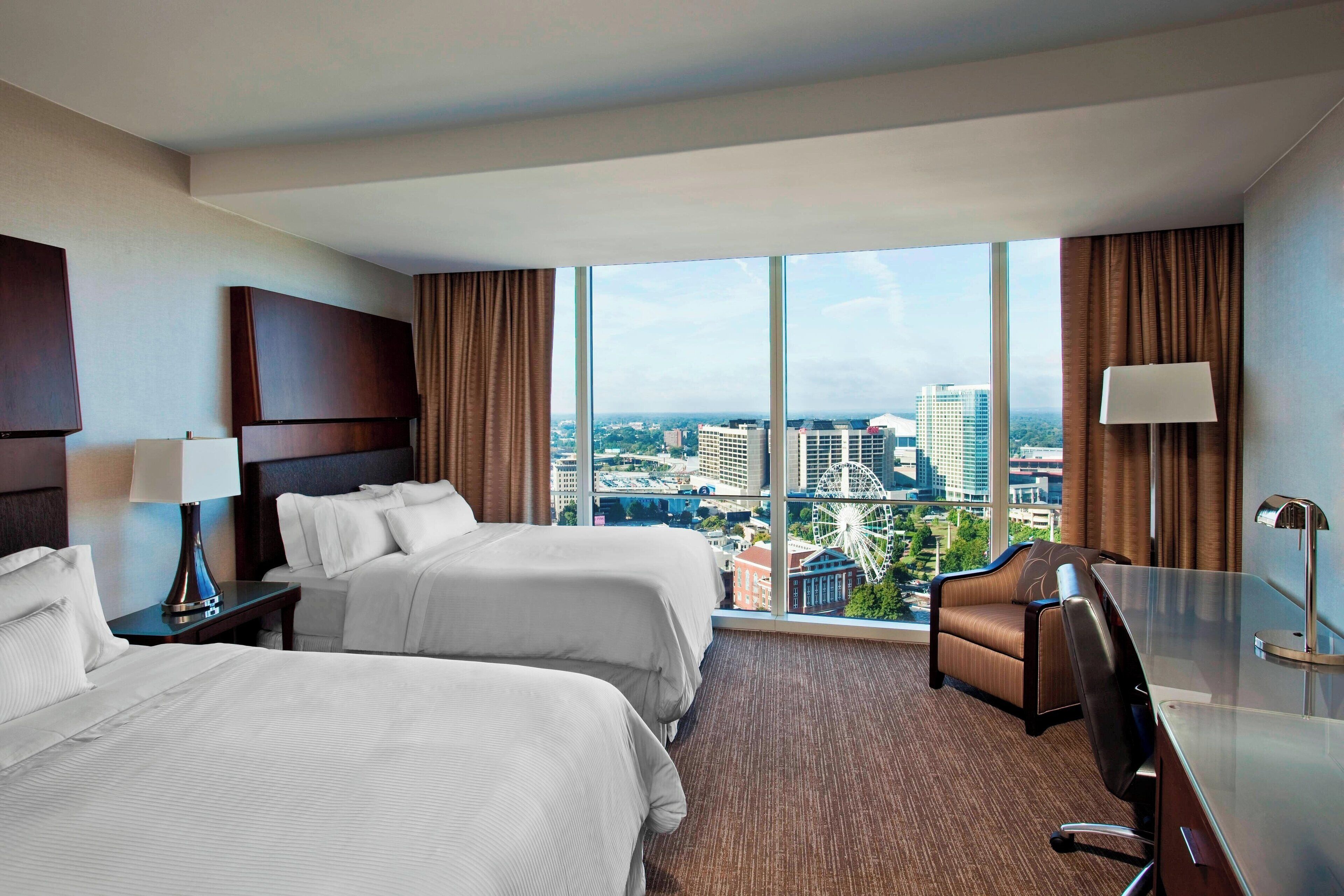A hotel room with two beds, a desk, a chair, and a large window offering a city view.