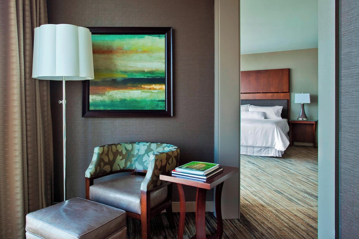 Een hotelkamer met een bed, een stoel, een tafel met boeken, een lamp en een schilderij aan de muur.