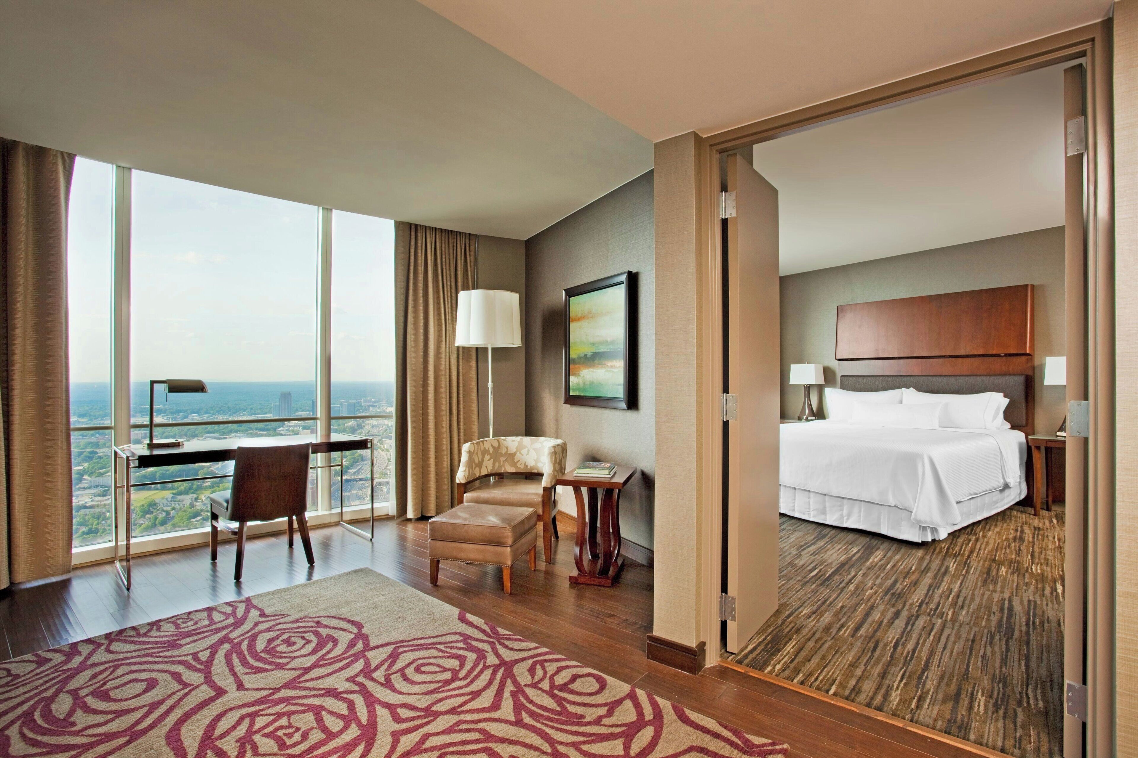 A hotel room with a large window, a desk, a chair, a bed, and a view of the cityscape.