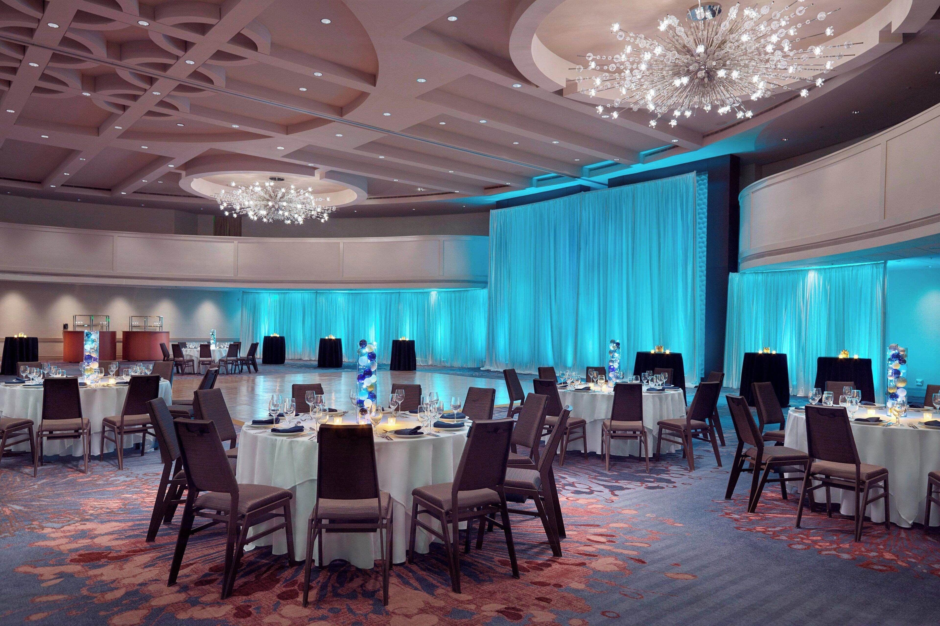 Un amplio salón de conferencias con mesas redondas, sillas y un fondo iluminado en azul.