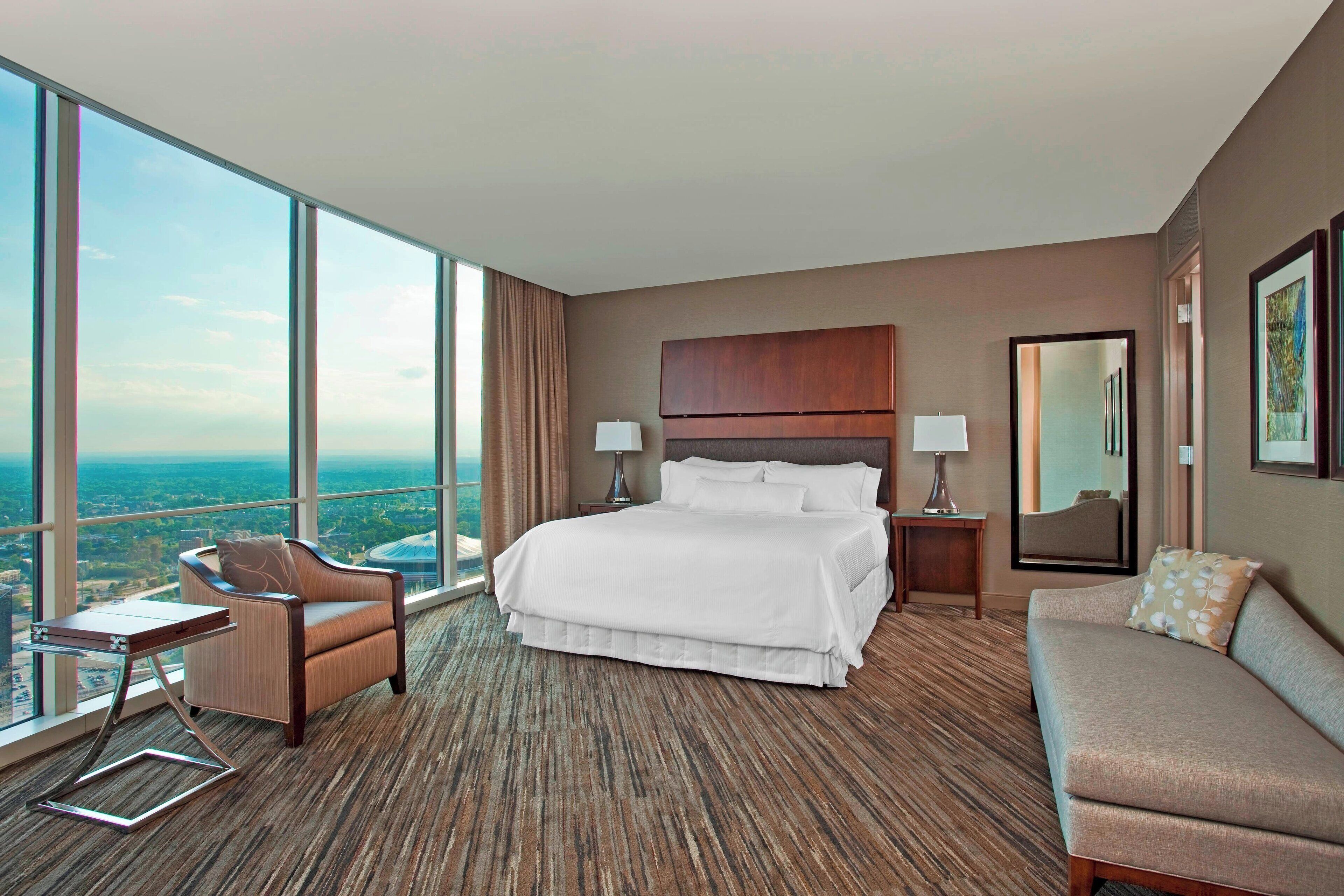 A hotel room with a large bed, a sofa, a chair, and a desk. There is a large window offering a city view.