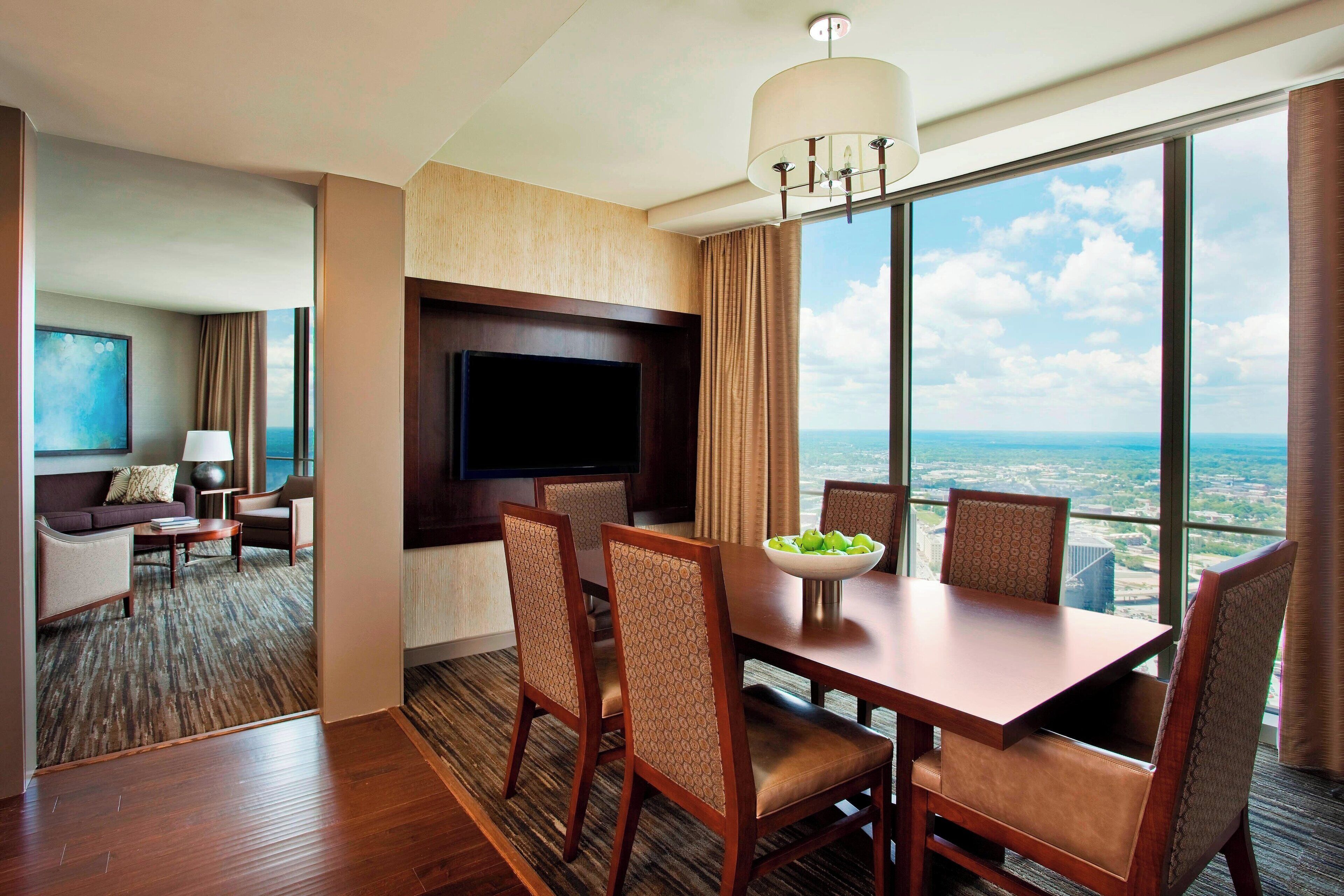 A hotel room with a dining area, a flat-screen TV, and a large window with a city view.