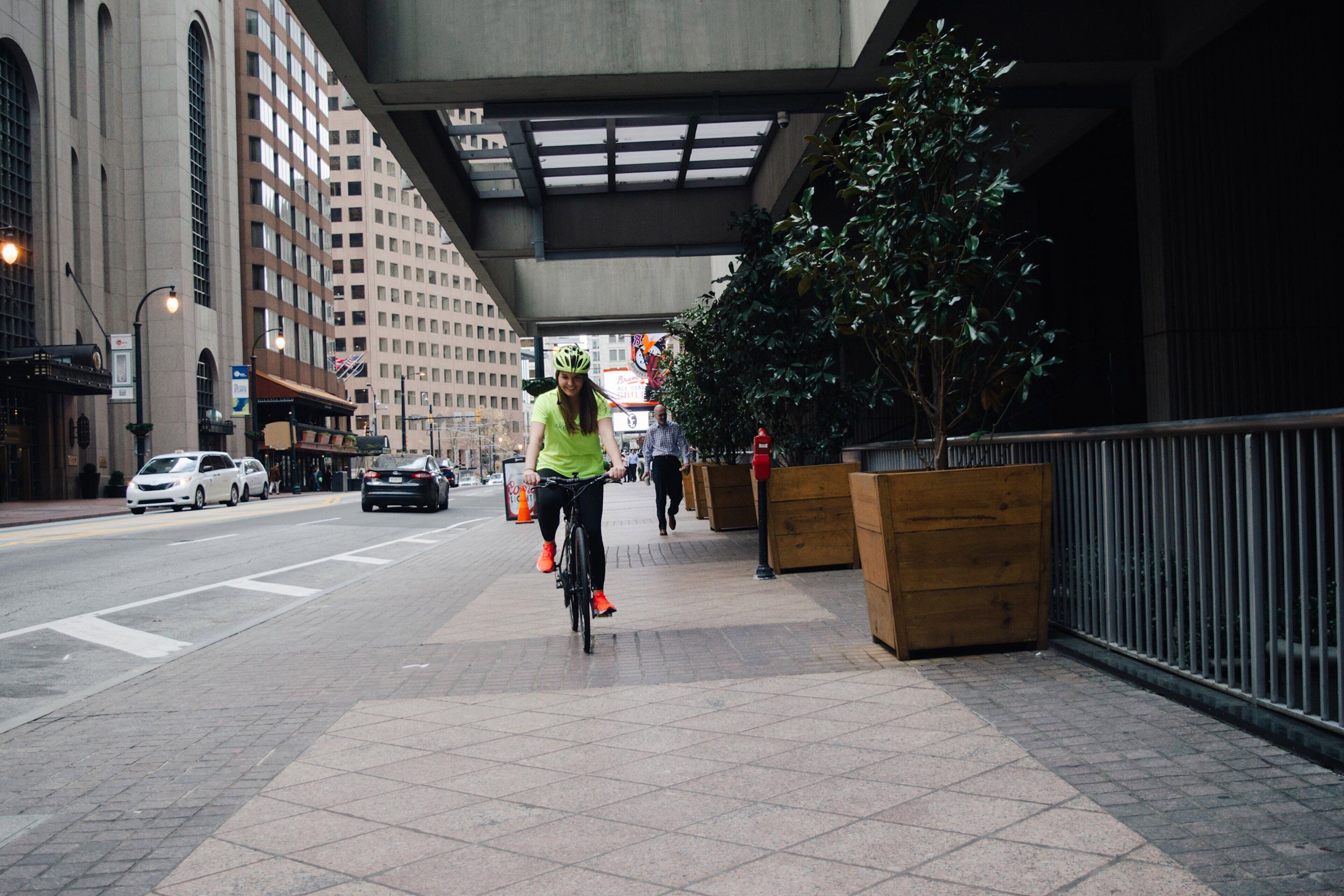 Un cycliste sur un trottoir en ville, avec des bâtiments et un arbre en arrière-plan.